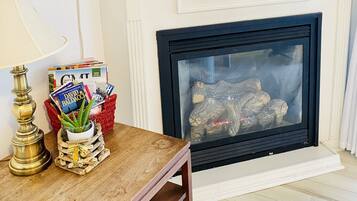 A living room with a flat-screen TV mounted above a white fireplace, a wooden table with magazines and a lamp, and a red basket with books and a plant.