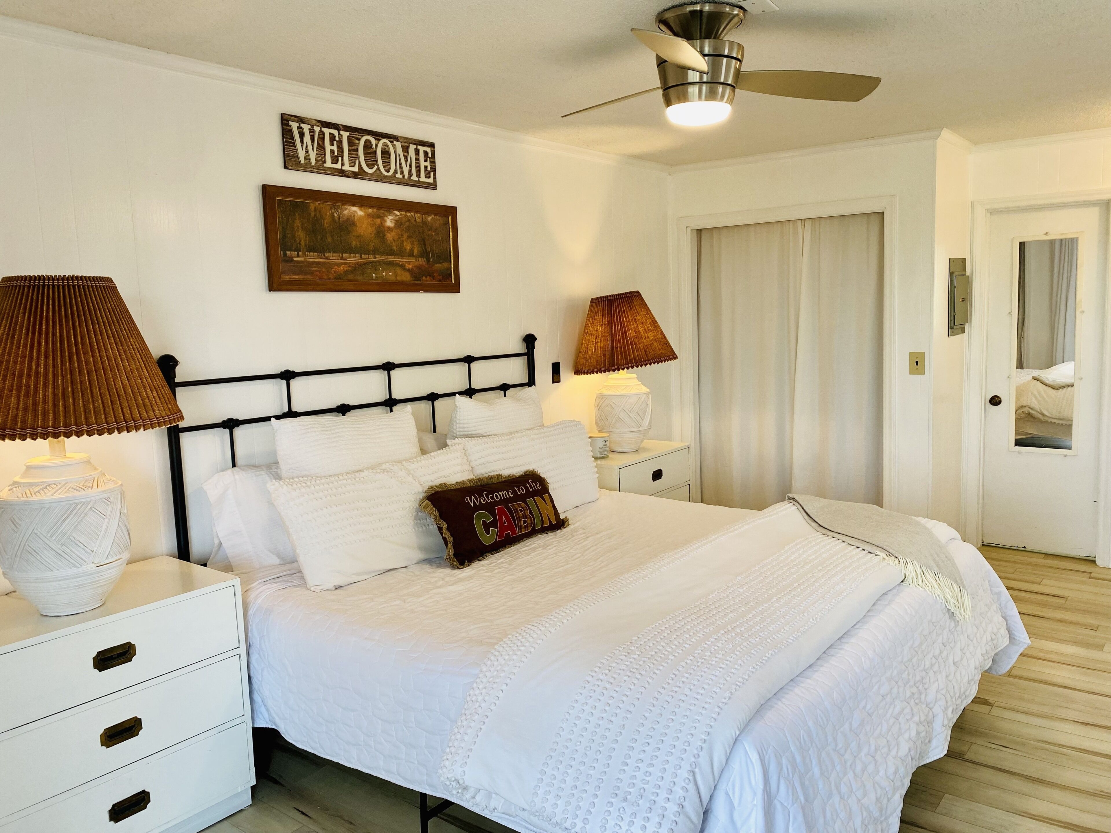 A neatly arranged bedroom with a bed, two bedside tables, a ceiling fan, a 'WELCOME' sign, and a door with a mirror.
