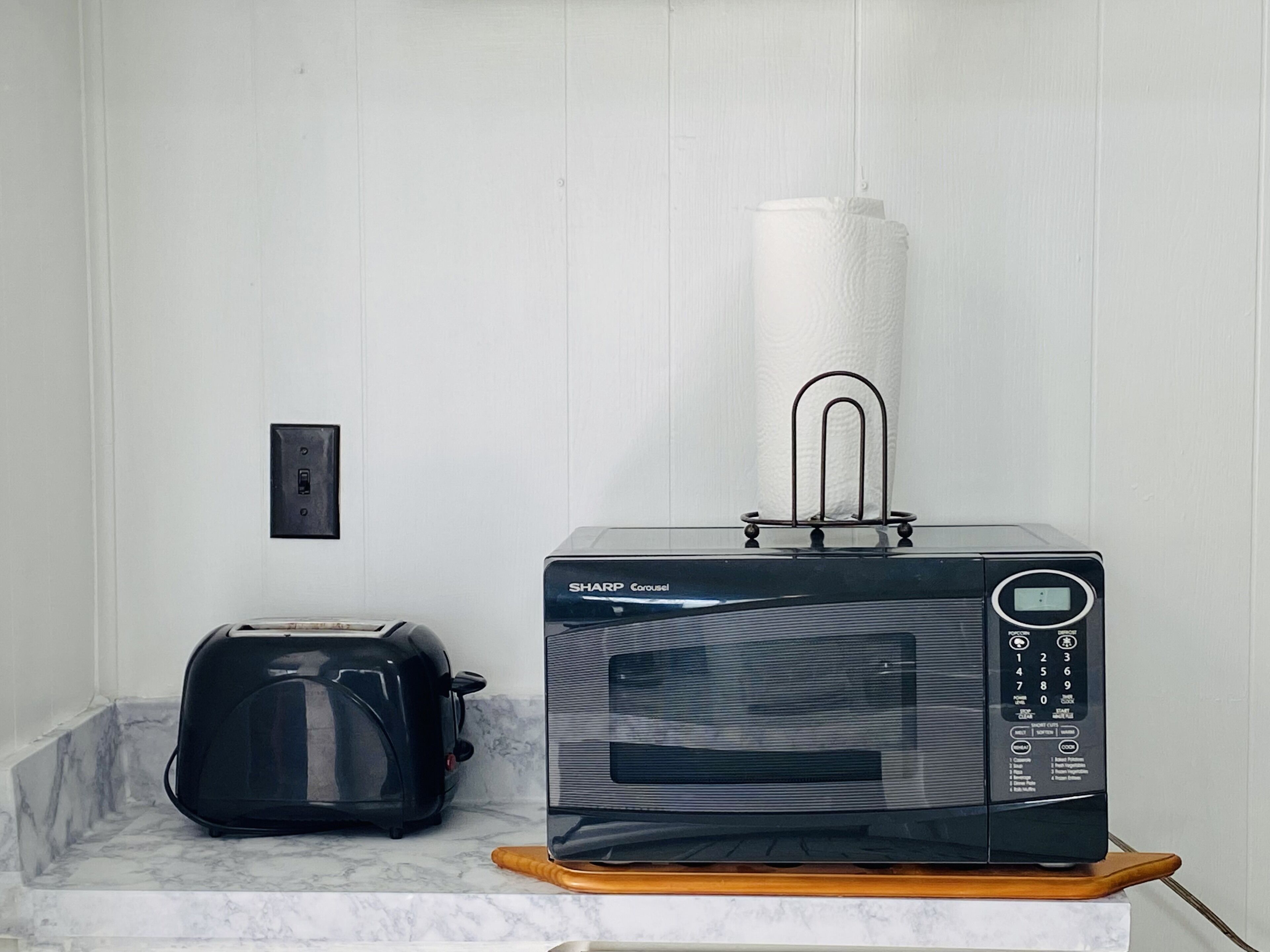 A microwave oven on a wooden stand with a paper towel holder on top, a toaster, and a power outlet on the wall.