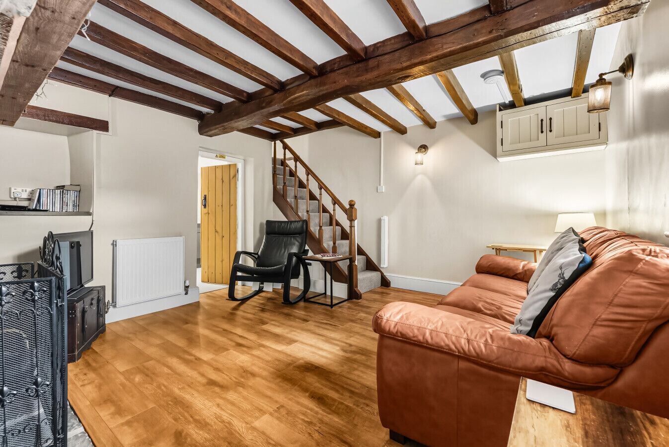 Uma sala de estar ampla com piso de madeira, um sofá de couro marrom, uma poltrona de balanço preta, uma escada de madeira e um radiador branco.