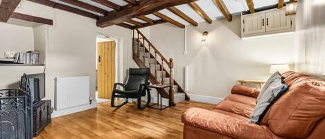 Uma sala de estar ampla com piso de madeira, um sofá de couro marrom, uma poltrona de balanço preta, uma escada de madeira e um radiador branco.