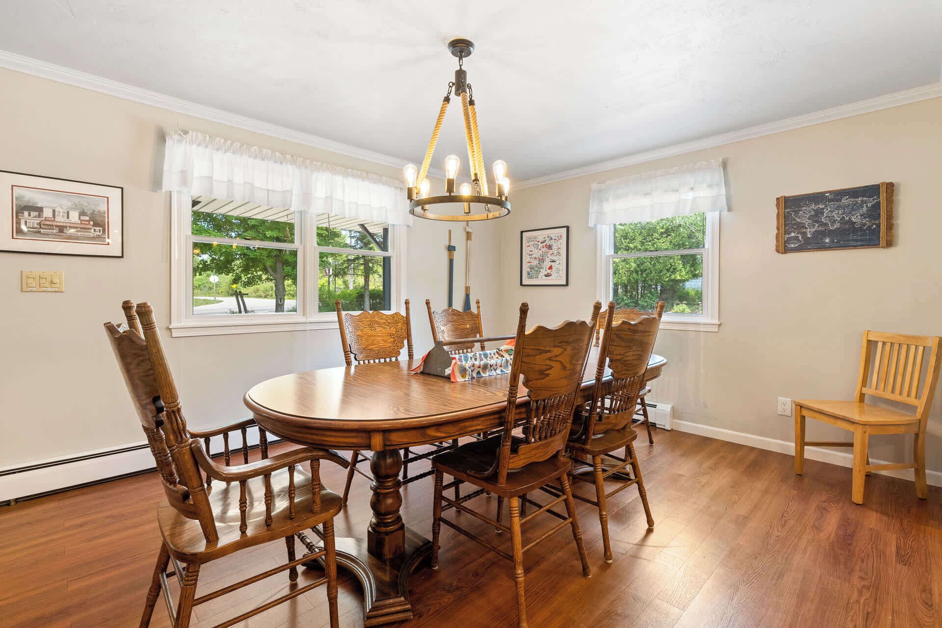 A dining room with a round wooden table, six chairs, a chandelier, and large windows with curtains.
