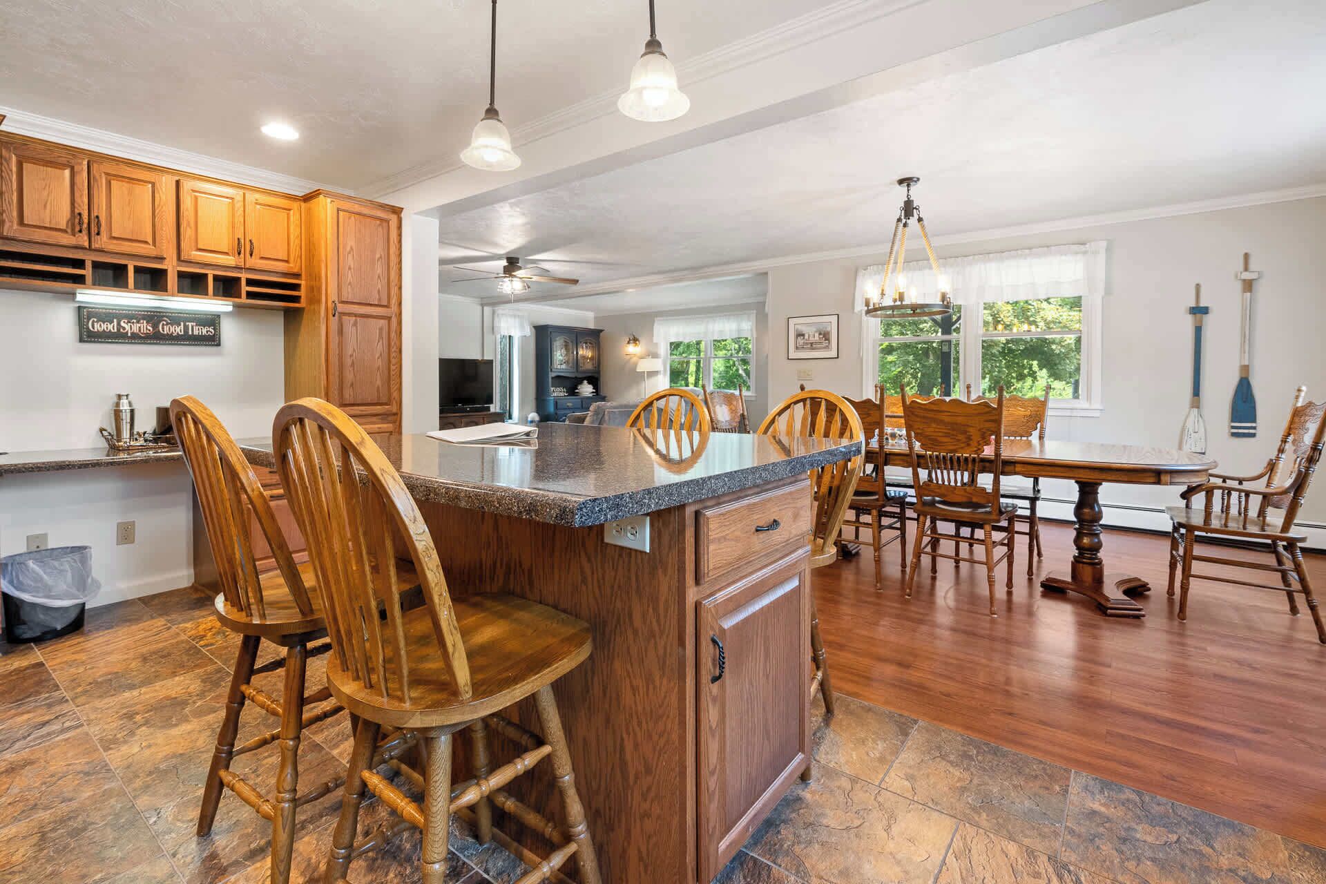 A spacious kitchen with wooden cabinets, a central island with bar stools, a dining area with a wooden table and chairs, and a view of the outdoors through large windows.