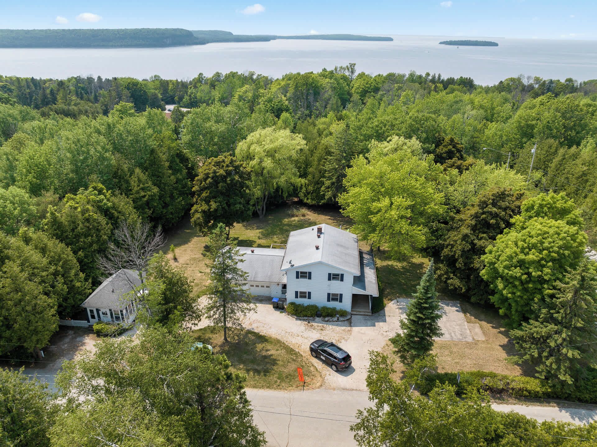 A white two-story house with a large front porch, surrounded by dense forest and a view of a lake.