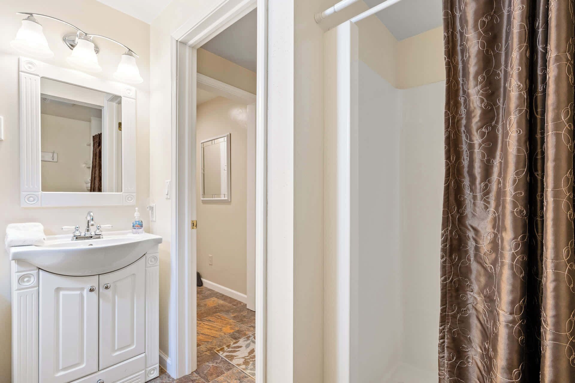 A bathroom with a white sink, mirror, and vanity, a shower curtain, and a hallway leading to another room.