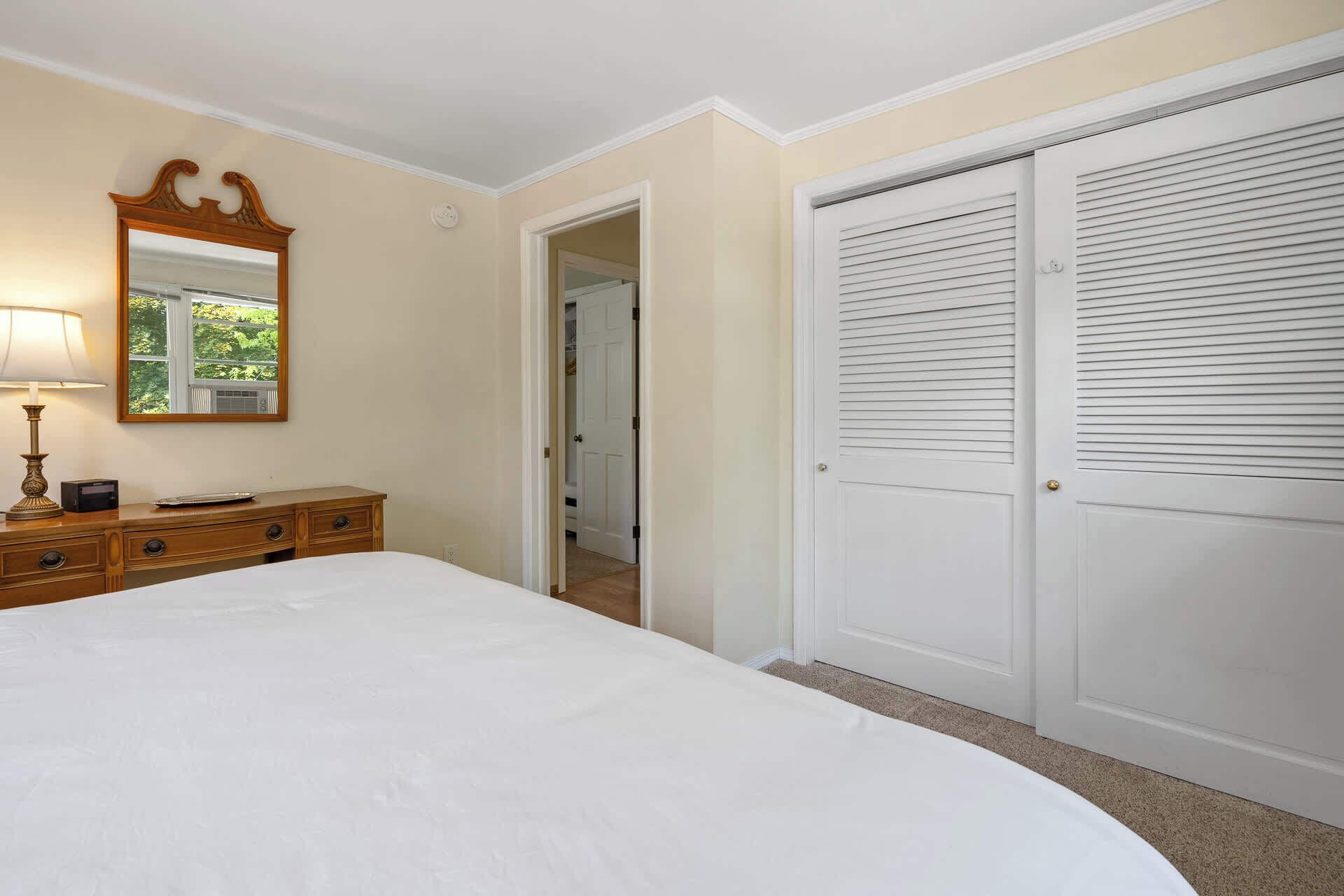 Quarto com cama, penteadeira, espelho e portas de guarda-roupa brancas.