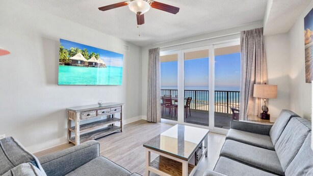 A spacious living room with a large window overlooking a balcony with a view of the sea. - Luxury 3BD Calypso Gulf Views! Free Beach Chairs Service for 4 (Panama City Beach)