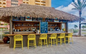 A bar with a thatched roof, yellow chairs, and a variety of bottles and glasses.
