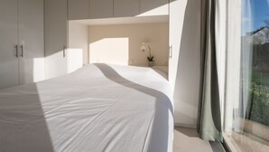 Une chambre avec un grand lit, une petite table sur laquelle est posée une plante, et une fenêtre avec des rideaux.