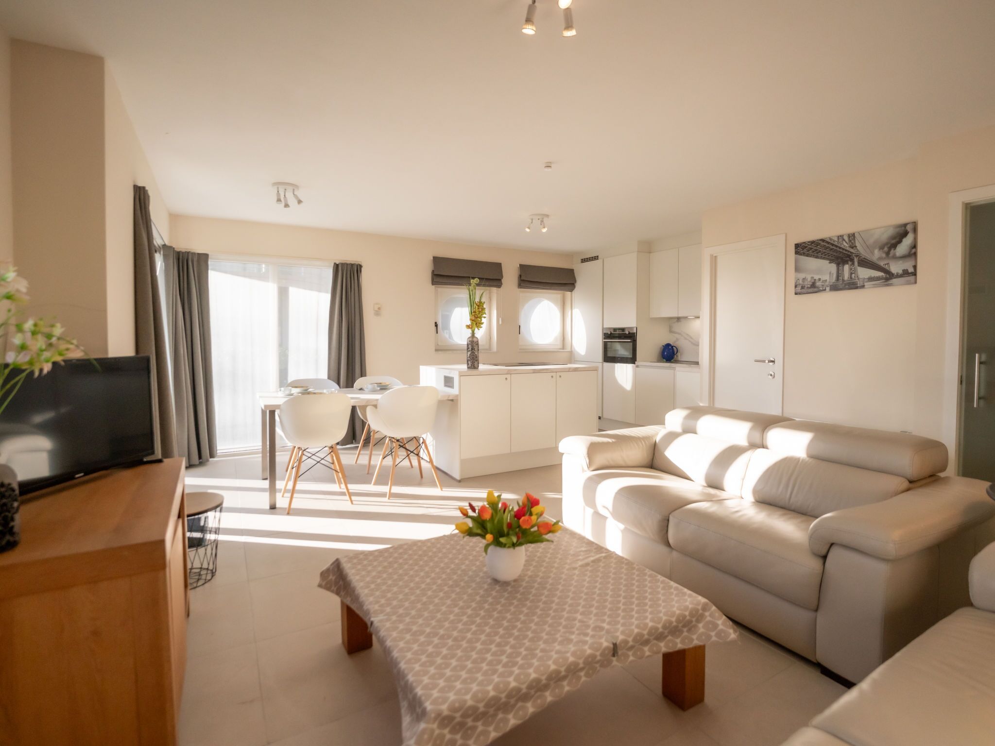 A modern living room with a sofa, a dining table with chairs, a TV, and a kitchen area in the background.
