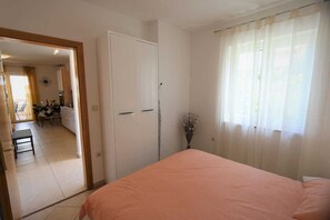 Quarto com cama, porta, janela com cortinas e vista para uma cozinha e sala de jantar.