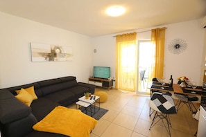 Un salon moderne avec un canapé noir, un téléviseur à écran plat, un coin repas comprenant une table et des chaises, et une porte-fenêtre coulissante donnant sur un balcon.