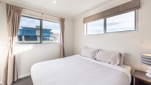 Une chambre à coucher avec un grand lit, deux fenêtres avec des rideaux, une vue sur un bâtiment à l’extérieur et une lampe de chevet.