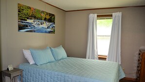 Un dormitorio con cama, mesita de noche, un cuadro y ventana con cortinas.