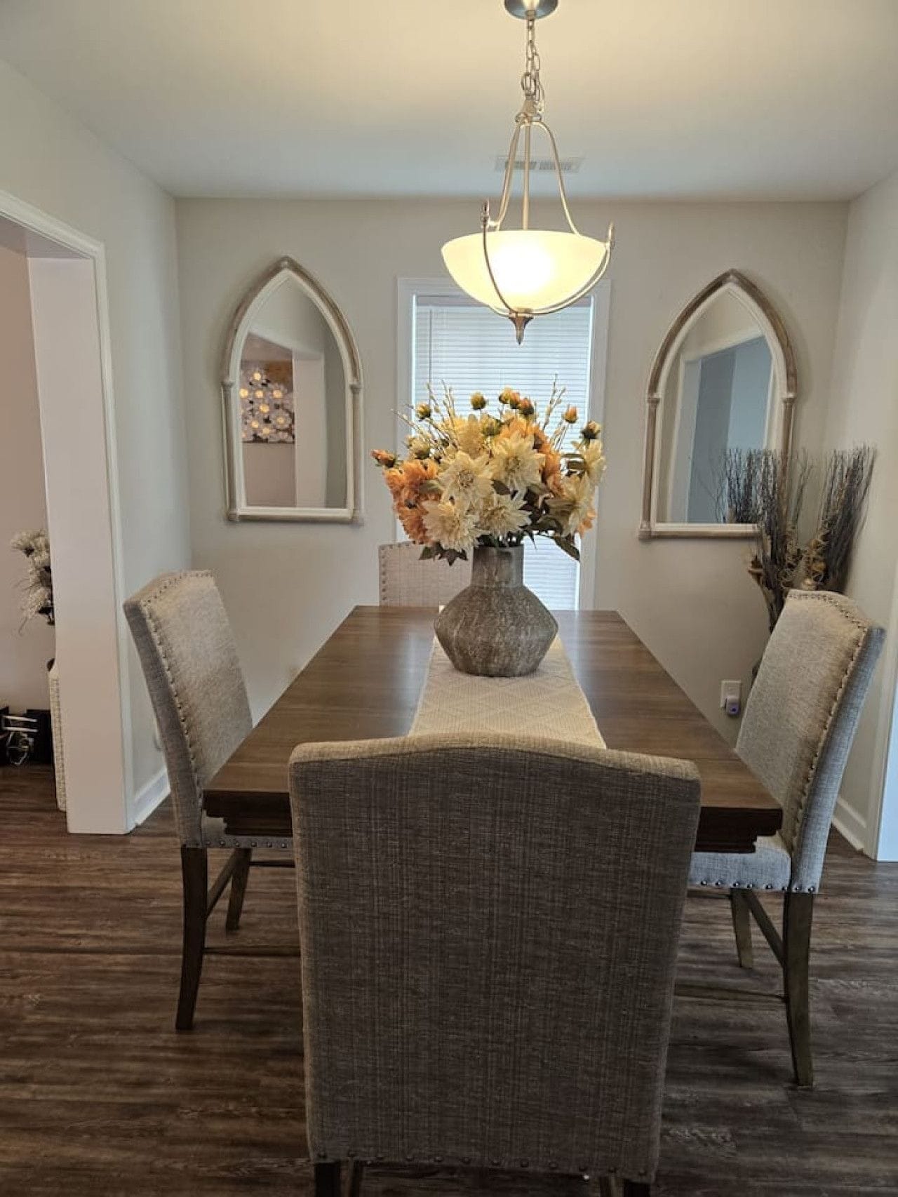 A dining room with a wooden table, four upholstered chairs, a vase with flowers, and two wall-mounted mirrors.