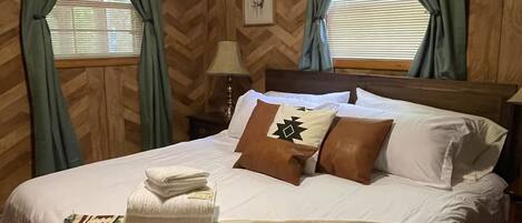 Een slaapkamer met een bed, kussens, een deken, een lamp en gordijnen.