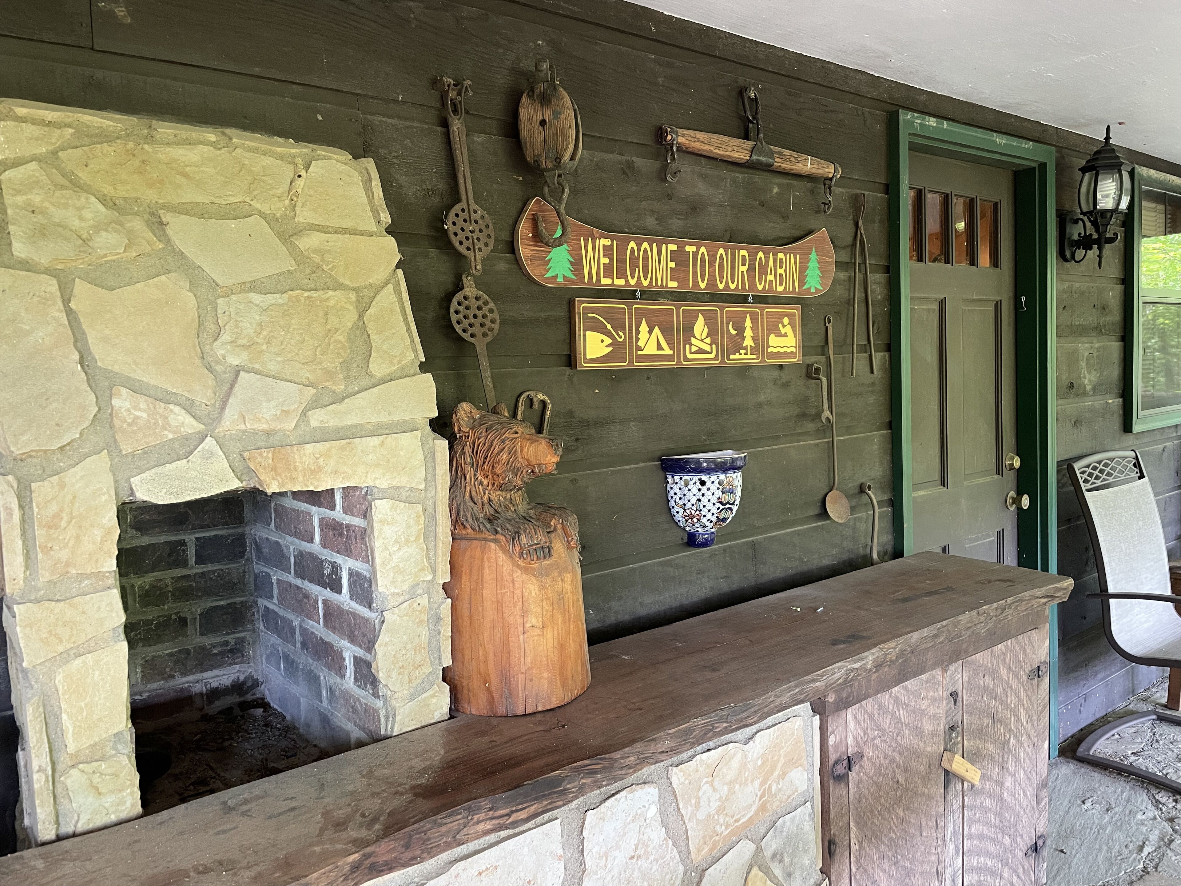 A cabin entrance with a stone fireplace, a wooden bear sculpture, a welcome sign, and a rustic door.