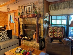 A cozy living room with a stone fireplace, wooden furniture, and a view of the outdoors. - Ken’s Secluded Cabin★Fire Pit★Huge Deck★Amazing Views★Hot tub★12 Minutes to DT (Bryson City)