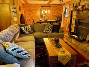 A cozy living room with a wooden herringbone floor, a sofa with patterned cushions, a coffee table with a fruit bowl, and a fireplace with decorative items. - Ken’s Secluded Cabin★Fire Pit★Huge Deck★Amazing Views★Hot tub★12 Minutes to DT (Bryson City)