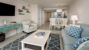 A living room with a sofa, armchair, coffee table, and a TV on the wall. There is a dining area with a table and chairs in the background. - Updated Beachfront Condo; Resort Amenities w/Heated Pool, Wifi & Beach Gear ~Walk to Dining & Shops (Marco Island)
