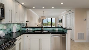 A modern kitchen with white cabinets, a black countertop, and a view of the outdoors. - Updated Beachfront Condo; Resort Amenities w/Heated Pool, Wifi & Beach Gear ~Walk to Dining & Shops (Marco Island)