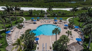 A large, circular swimming pool surrounded by lounge areas with umbrellas, palm trees, and a view of a river. - Updated Beachfront Condo; Resort Amenities w/Heated Pool, Wifi & Beach Gear ~Walk to Dining & Shops (Marco Island)