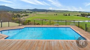 Una piscina rettangolare con bordo in legno e ringhiera in metallo nero, immersa in un prato e delimitata da una recinzione, con vista su colline lontane e cielo sereno.