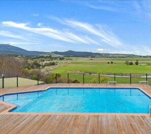 Une piscine rectangulaire dotée d’une terrasse en bois et d’une rambarde en métal noir, entourée d’une pelouse et d’une clôture, et offrant une vue sur des collines lointaines et un ciel dégagé.