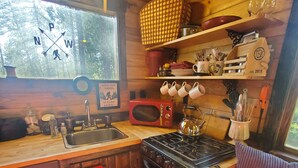 A compact kitchen with wooden cabinets, a microwave, a stove, and a sink. There are hanging mugs and a shelf with kitchen utensils.