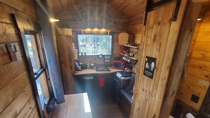 A compact wooden cabin with a kitchenette, a small dining table, and a view of the outdoors through a window.