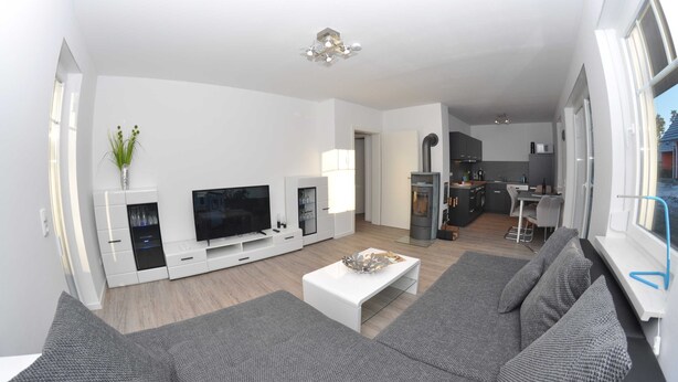 A modern living room with a grey sofa, a white coffee table, and a TV on a white entertainment unit. There is a kitchen area visible in the background. - 3 Zimmer Erdgeschoss - Fewo Ferienhaus Müritzglück - Ferienwohnungen / Appartements - Ferienhaus (Röbel/Müritz)