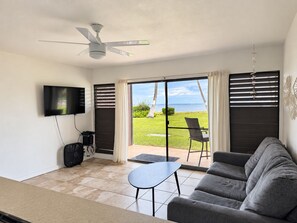 A living room with a flat-screen TV, ceiling fan, and a view of the ocean through large sliding doors. - Beautifully Renovated Beachfront Condo (Kaunakakai)