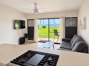 A modern living room with a flat-screen TV, a ceiling fan, a sofa, and a view of the ocean through large sliding doors. - Beautifully Renovated Beachfront Condo (Kaunakakai)