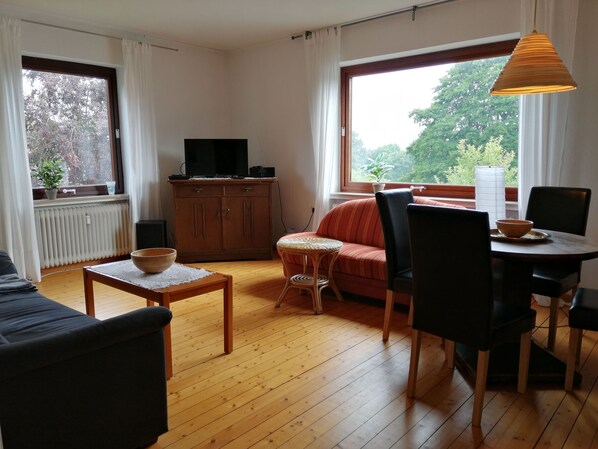 A living room with a sofa, a dining table with chairs, a television on a cabinet, and a window with a view of trees.