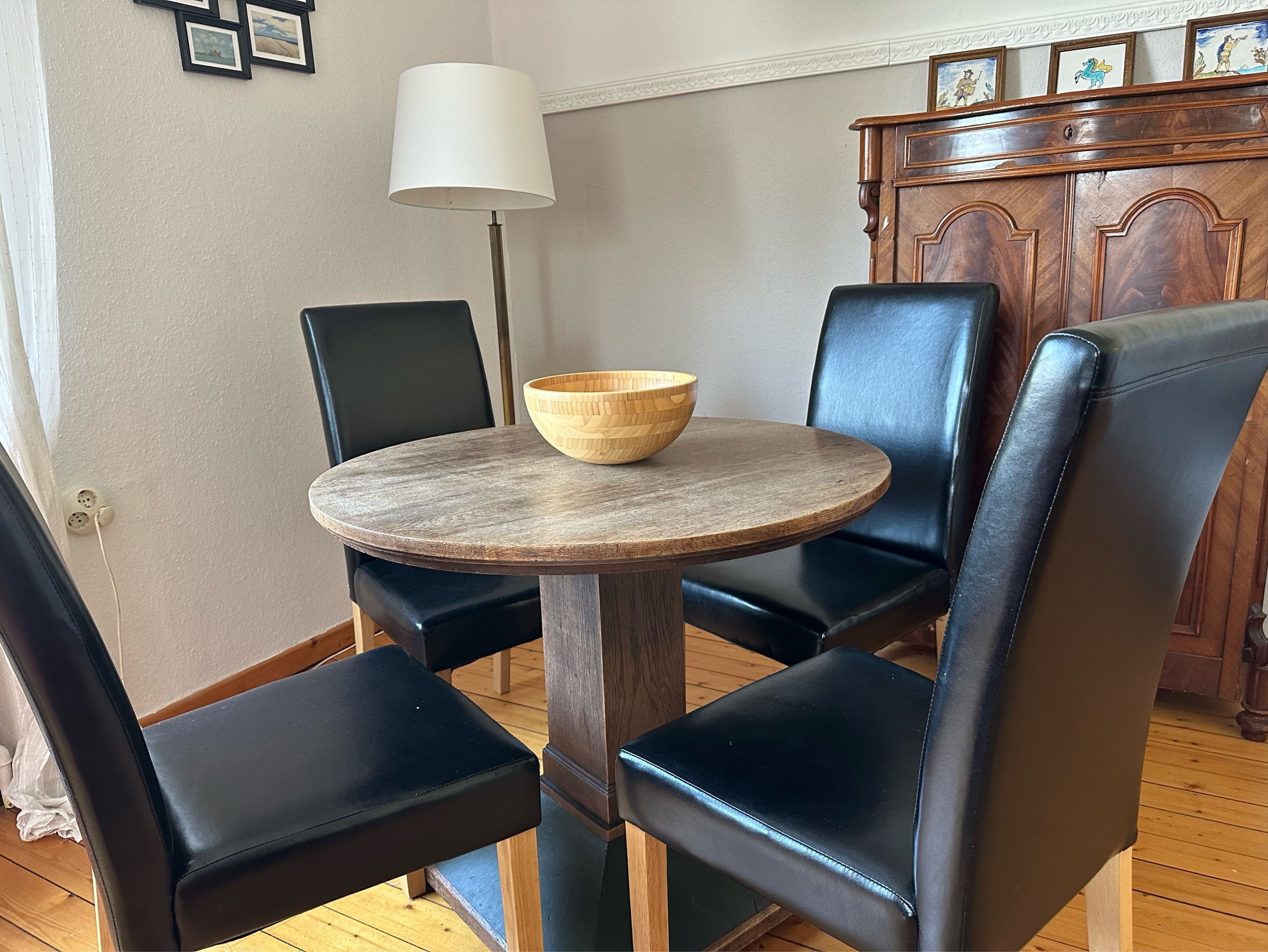 Un comedor con una mesa redonda de madera, cuatro sillas negras, un cuenco de madera, una lámpara de pie y un mueble de madera con obras de arte.