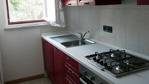 A kitchen with red cabinets, a white countertop, a sink, a stove, and a window with a view of trees. - Maison Perchu - Pila, Vallée D'aoste (Pila)