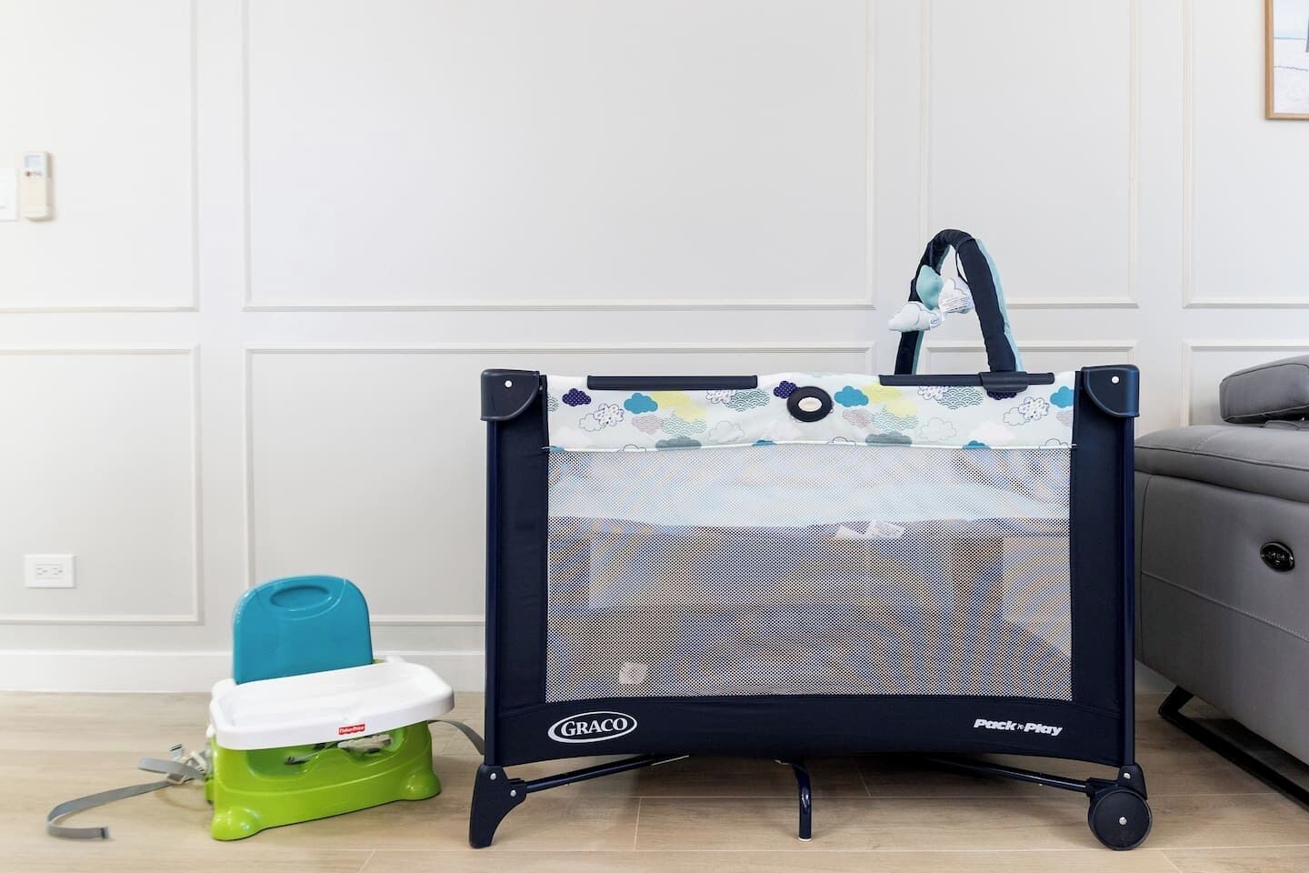 A Graco Pack n Play with a baby seat, a green potty, and a grey sofa in a room with white walls and a wooden floor.