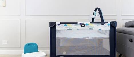 A Graco Pack n Play with a baby seat, a green potty, and a grey sofa in a room with white walls and a wooden floor.