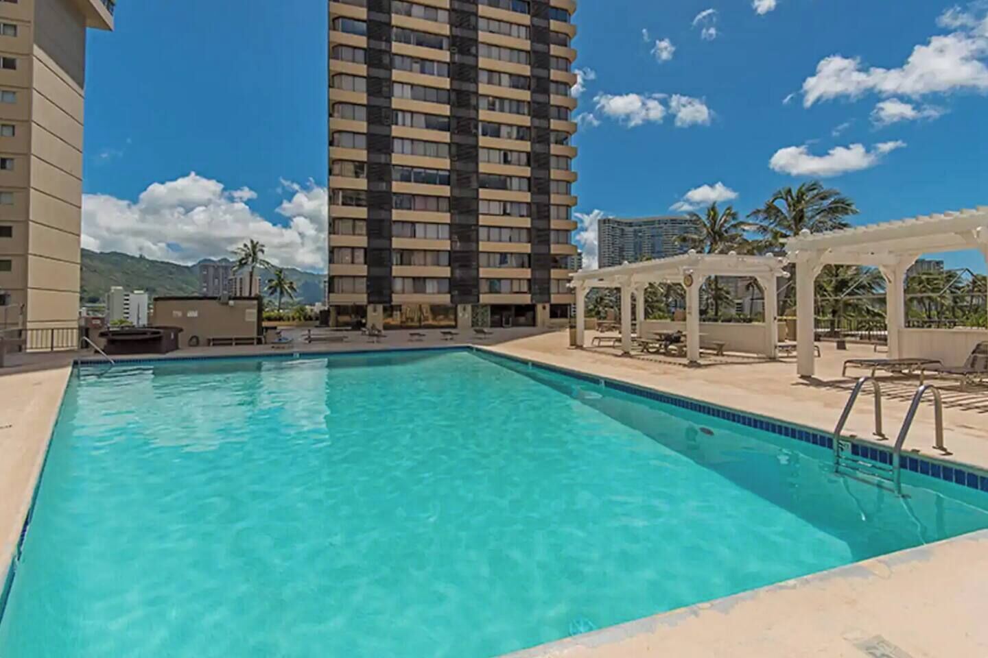 Una piscina con agua cristalina azul, rodeada por un área de descanso con mesas y sillas, y un edificio alto al fondo.