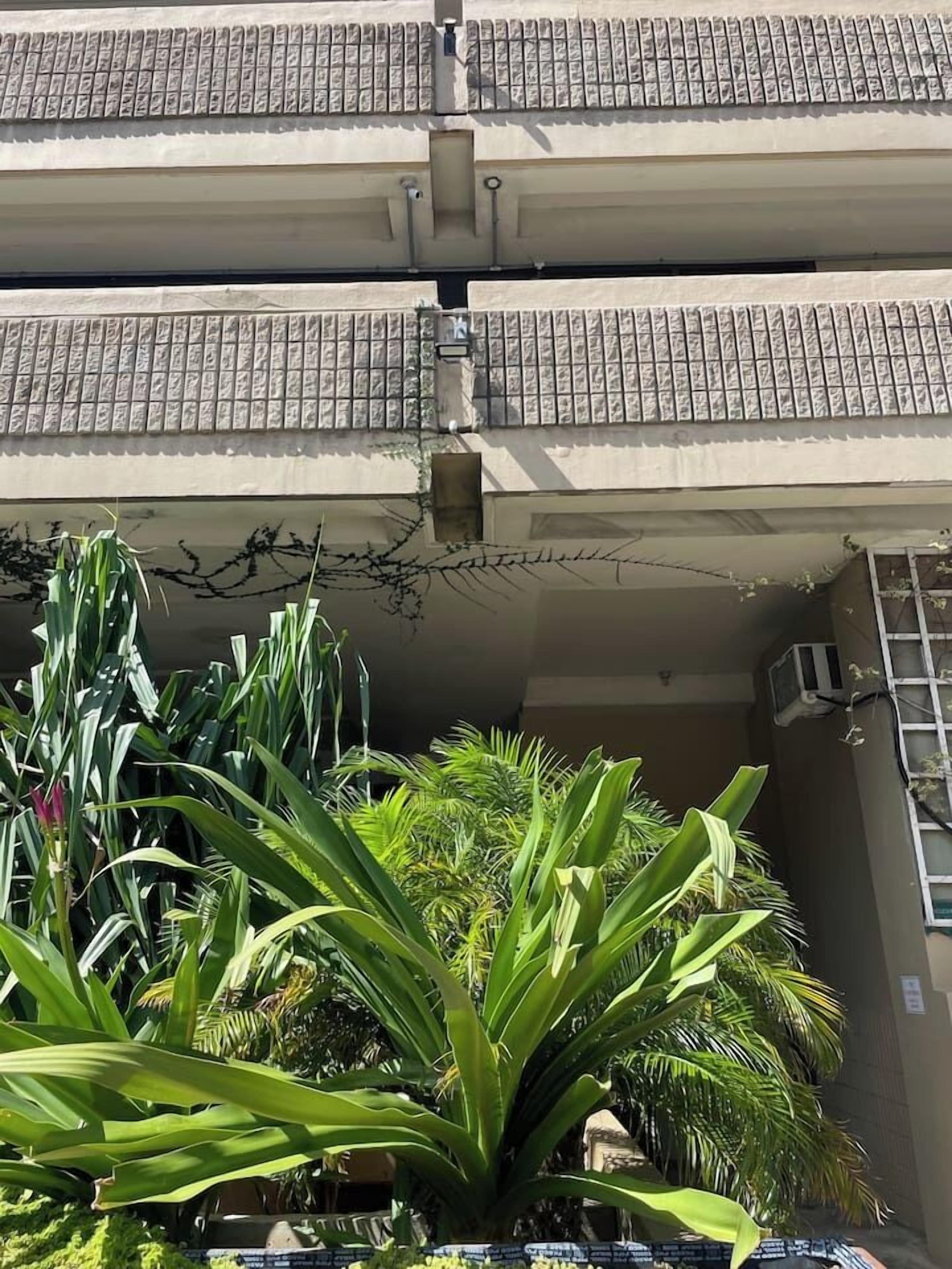 A multi-story building with a tiled roof, a visible air conditioning unit, and lush green plants in the foreground.
