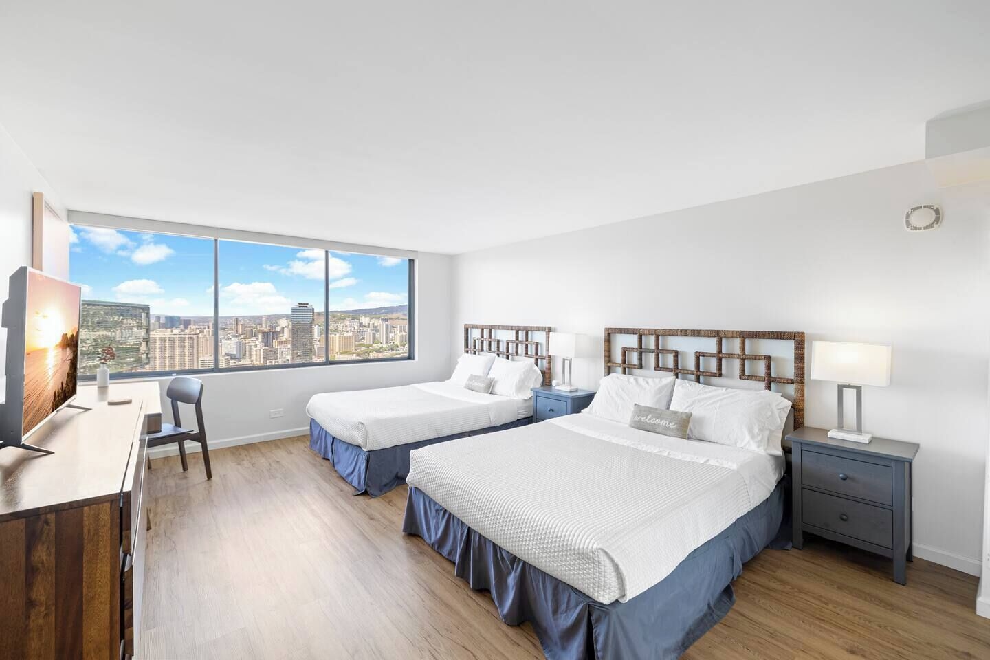 A hotel room with two beds, a desk, a chair, and a large window with a city view.