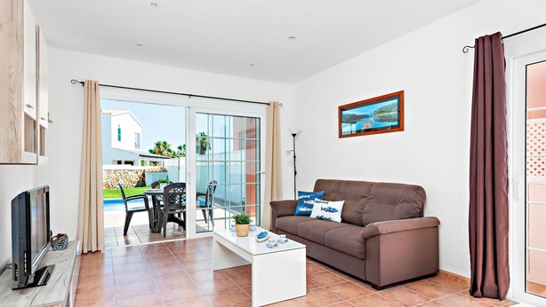 A modern living room with a sofa, coffee table, and a TV. There is a glass door leading to an outdoor area with a pool and chairs.
