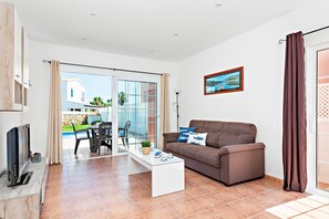 Uma sala de estar moderna com sofá, mesa de centro e televisão. Há uma porta de vidro que dá para área externa com piscina e cadeiras.