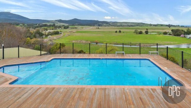 Piscina retangular com deck de madeira e guarda-corpo de metal preto, cercada por gramado e cerca, com vista para colinas distantes e céu claro.