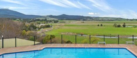 Une piscine rectangulaire dotée d’une terrasse en bois et d’une rambarde en métal noir, entourée d’une pelouse et d’une clôture, et offrant une vue sur des collines lointaines et un ciel dégagé.