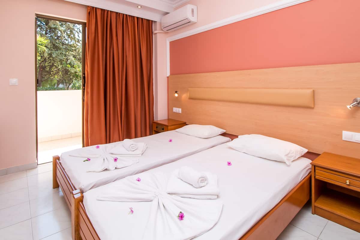 Une chambre d’hôtel avec une tête de lit en bois, deux lits avec du linge de lit blanc et des pétales de fleurs roses, une table de chevet en bois et un climatiseur fixé au mur.