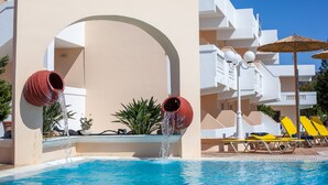 Un espace piscine doté de deux jets d’eau, d’un bâtiment blanc et de chaises longues jaunes.