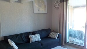 Un sofà de color fosc amb coixinets estampats, una taula de cafè blanca, un rellotge de paret i una finestra amb cortines.