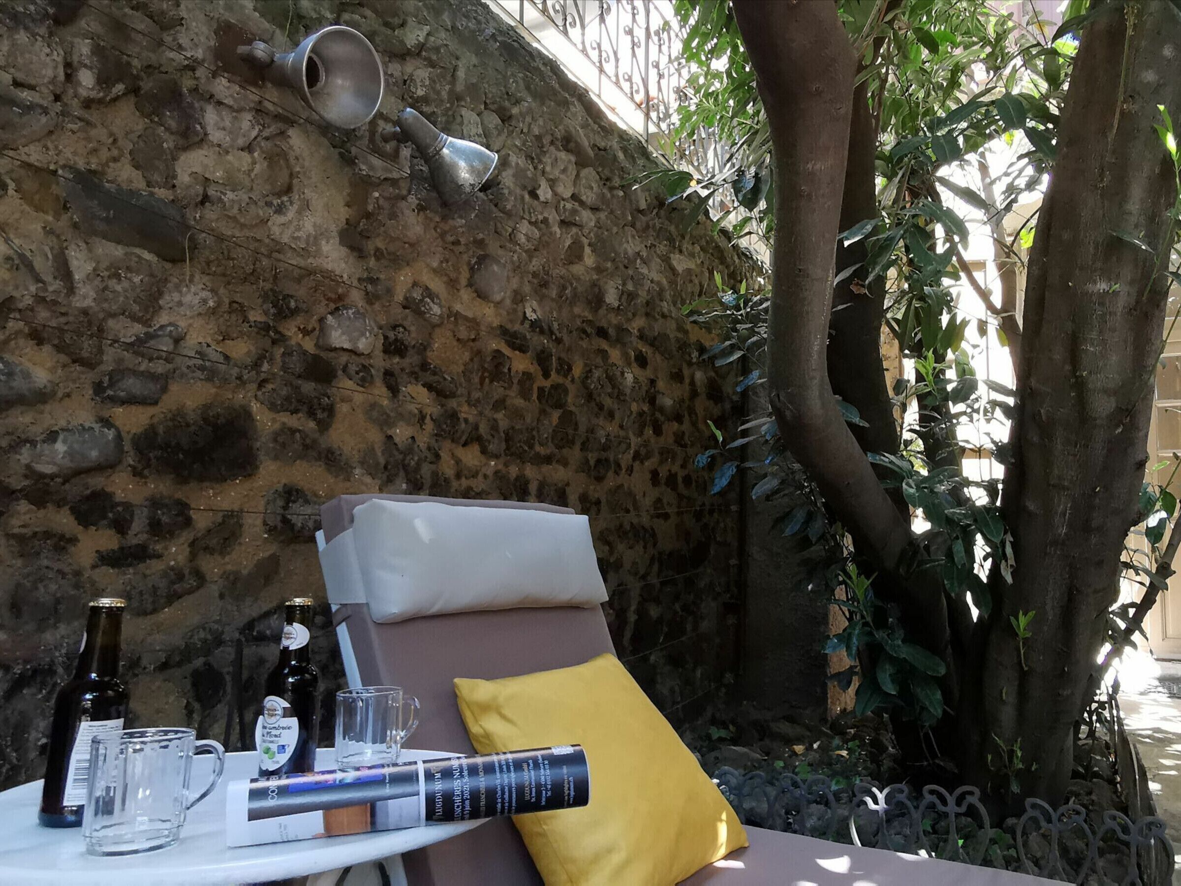 A stone wall, a metal bell, a tree, and a table with a cushion and a yellow pillow.