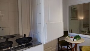 Una cocina con estufa, dos sartenes, un lavamanos y una ventana que ofrece vistas al exterior. Hay una mesa pequeña con una silla y una planta encima.
